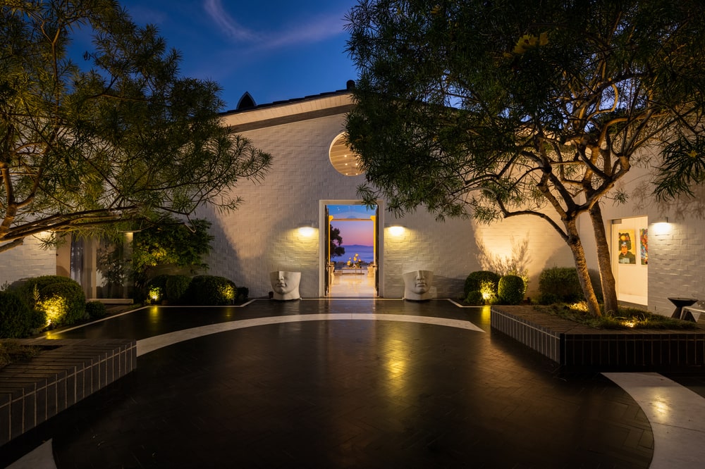 This is a nighttime view of the small courtyard of the entrance of the house with a shiny dark floor and warm lighting on the landscaping. Image courtesy of Toptenrealestatedeals.com.