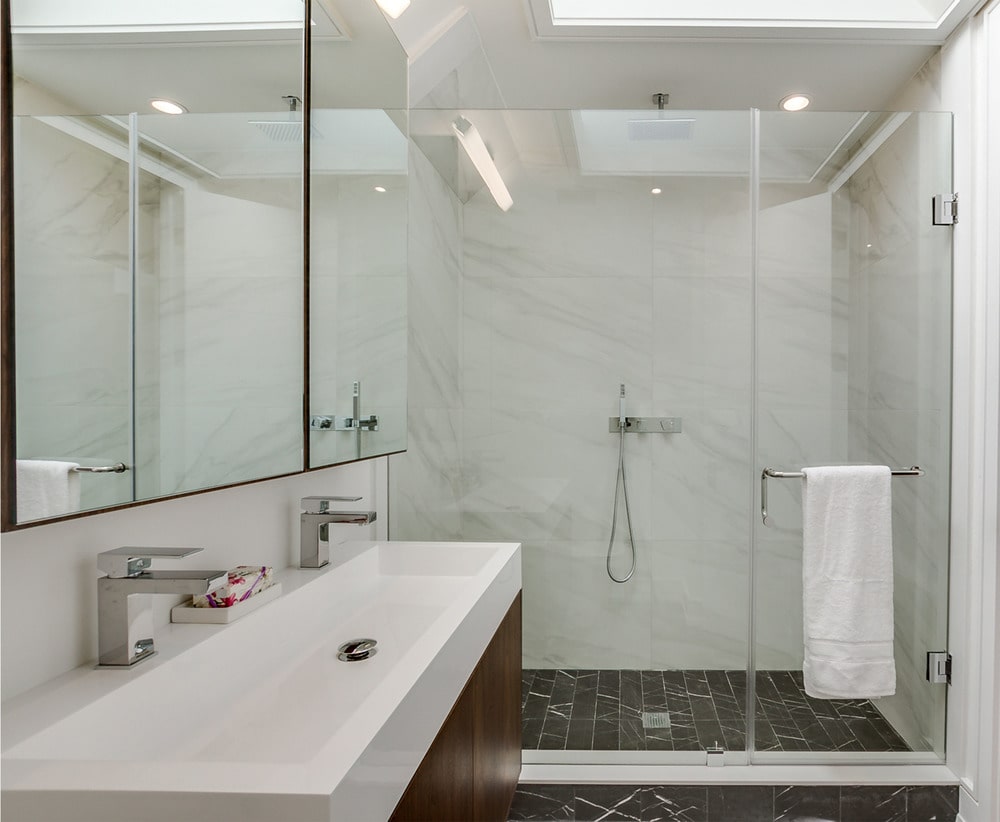 The bathroom has a glass-enclosed shower area on the far side with dark tiles on the floor and white marble tiles on the walls.