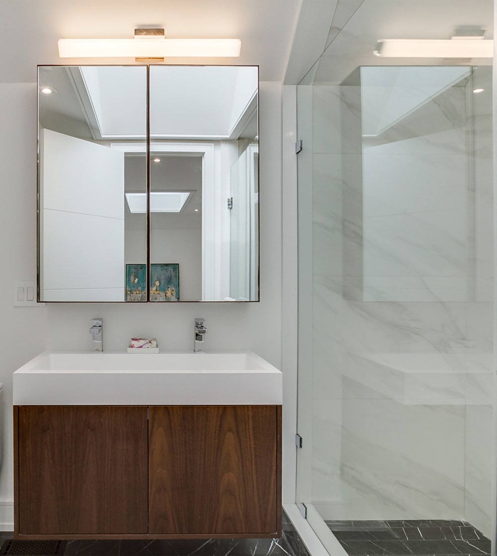 On the side of this shower area is the vanity that has a white sink and counter contrasted by the dark wooden cabinet below and the mirror above.