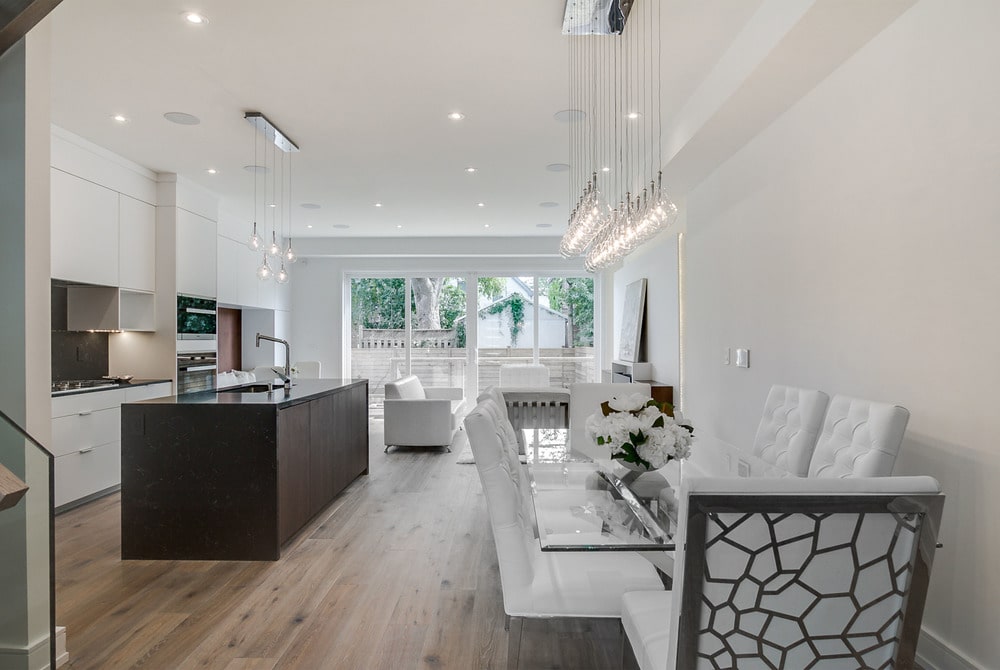 This is a view of the kitchen from the vantage of the dining area that has a large dark wooden kitchen island that is contrasted by the surrounding white structures.