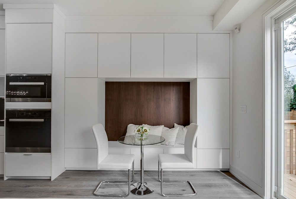 This is a close look at the breakfast nook just beside the kitchen with a small round glass-top dining table paired with white modern chairs and a built-in alcove seating.