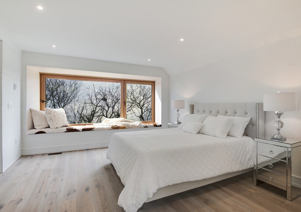 This bedroom has a large white bed and a reading nook below the window on the far side with a built-in bench under the glass windows.