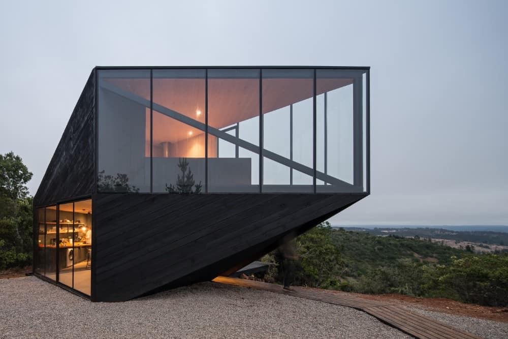 This is a closer look at the exterior of the house with black wooden shiplap exterior walls and glass walls on both levels showcasing the interiors with a warm glow.