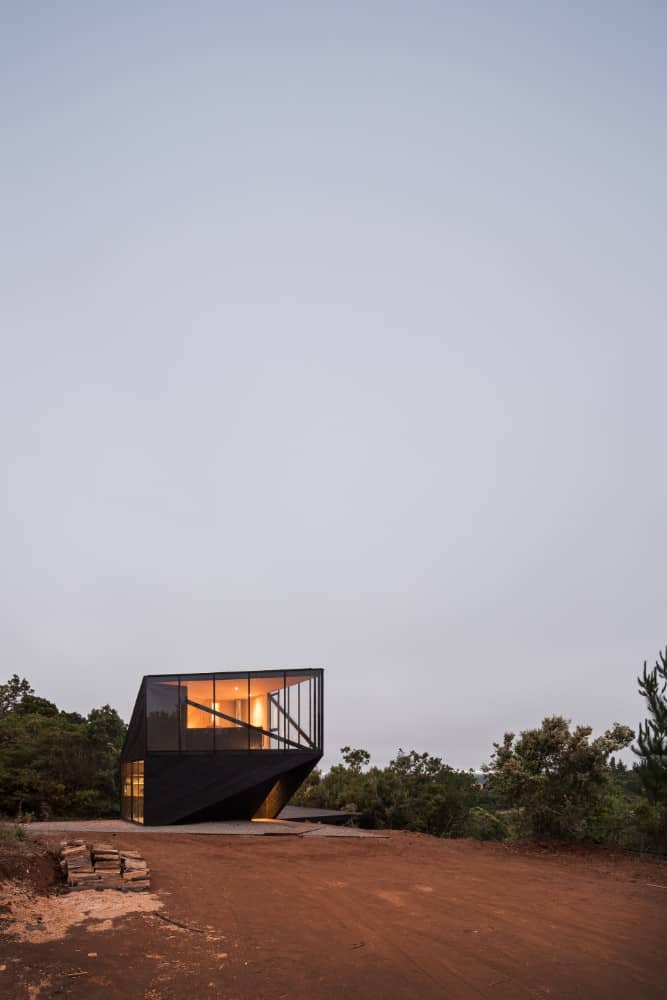 This is a view of the modern house with dark walls and warm lighting that makes it stand out against the surrounding landscape of trees and soil.