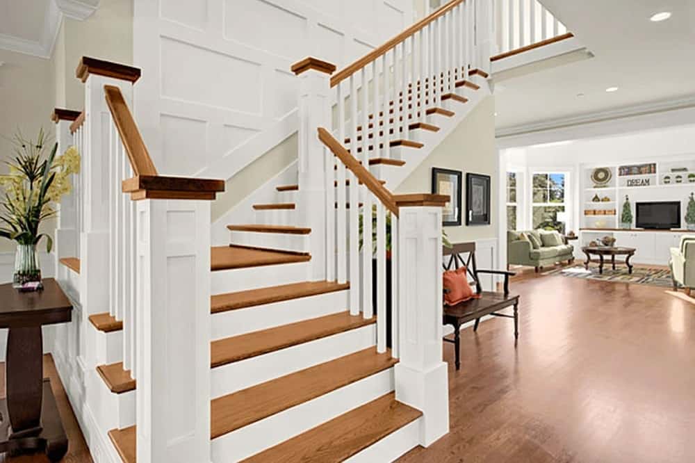 This is a close look at the foyer that has a large wooden staircase that has wooden steps that match the hardwood flooring. On the side of this is a brown bench with a throw pillow and wall artworks.