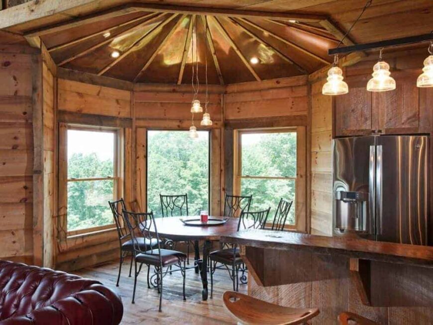 This dining area beside the kitchen and living room has large windows that brighten the dark round table surrounded by wrought-iron dining chairs topped with an arched ceiling.