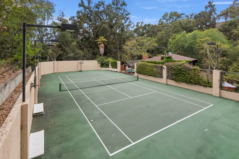 The tennis court is surrounded by tall concrete walls and adorned by the shrubs and the tall trees in the background. Image courtesy of Toptenrealestatedeals.com