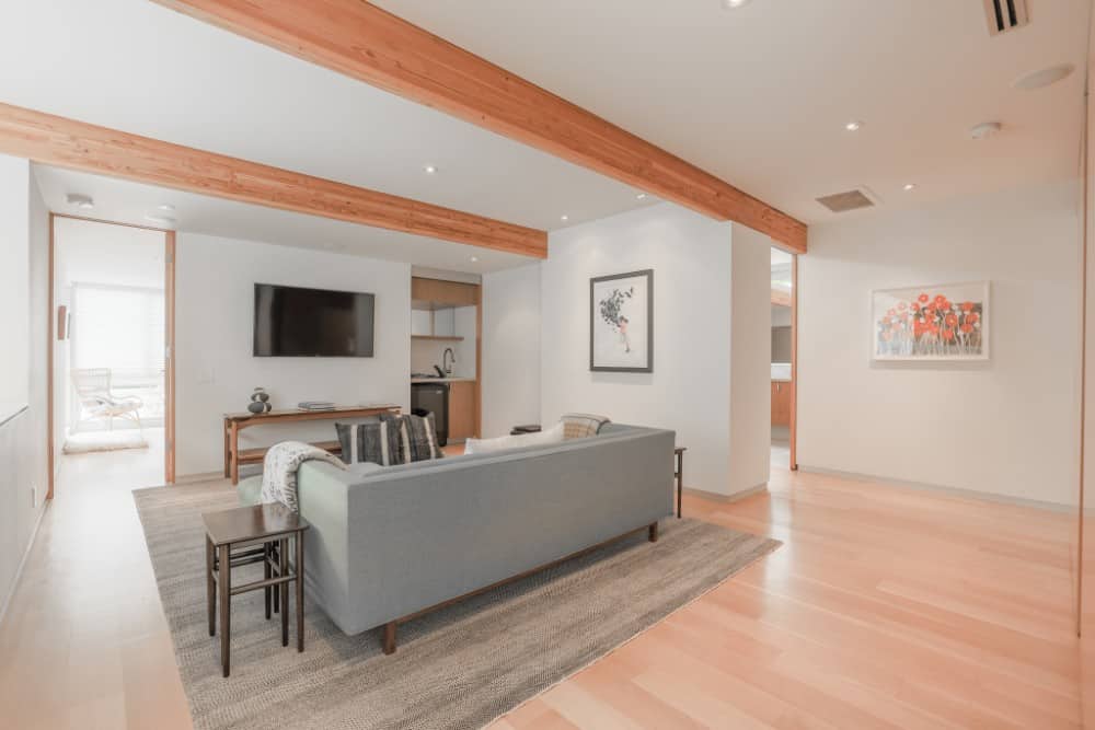 The beamed ceiling continues on the family room filled with a gray sofa, dark wood tables, and a wall-mounted TV. Image courtesy of Toptenrealestatedeals.com.