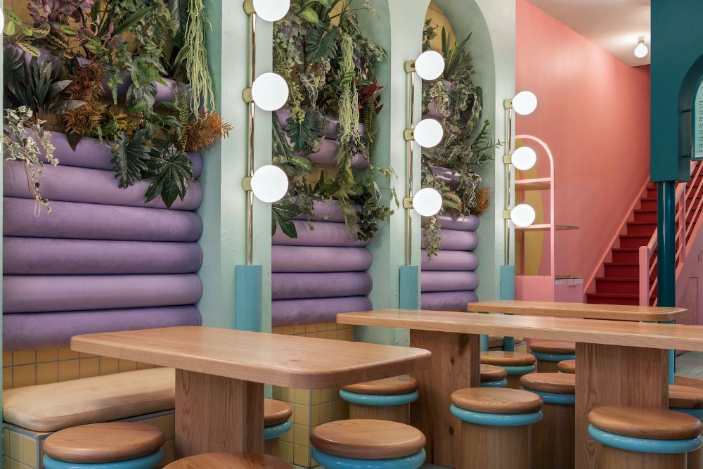 Restaurant dining space with wooden rectangular tables, wooden stool-styled chairs, arc side wall design with custom lighting and plants, and a wooden stair leading to the second story.