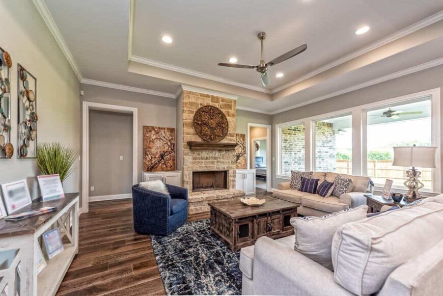 The family room has beige sofas, a stone fireplace, and a trio of windows that bring an abundant amount of natural light in.