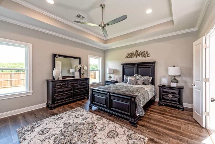 The primary bedroom has dark wood furnishings and a tray ceiling mounted with a chrome fan.