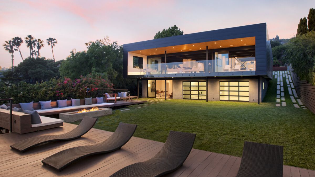 The exterior of the house showcases a stunning architectural design, characterized by its ultra-modern aesthetic. The ample driveway and lawn area, as well as the presence of sun loungers and a fireplace, highlight the property's outdoor living spaces and create a comfortable and inviting atmosphere.