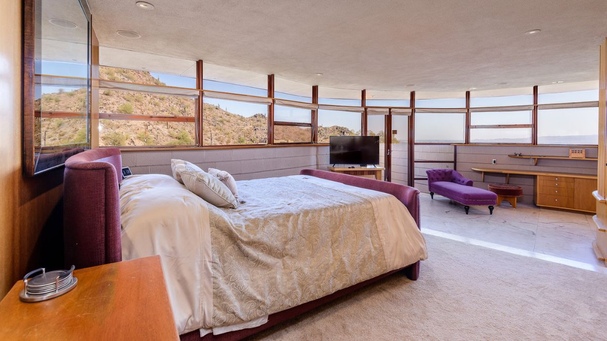 The master bedroom is carpeted and spotless, with a wooden side table, TV set, and chair that matches the color of the bed frame, and its window provides light and a view of the mountains.