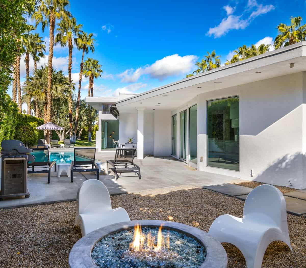 The backyard is filled with a swimming pool and a fire pit paired with white contemporary chairs.