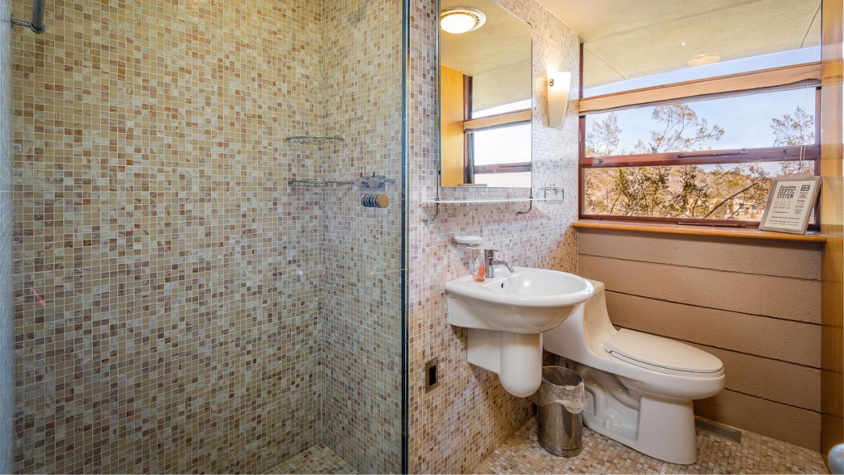 The comfort room has a square-shaped mirror, a tiny white sink, a rectangular window, and a white toilet with a tile and wooden wall combination.