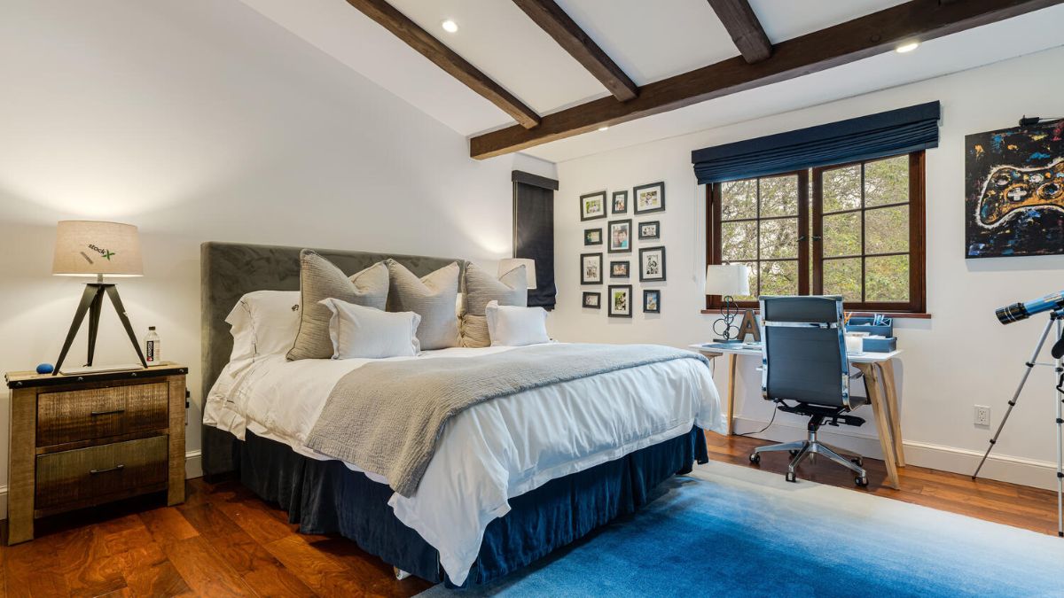 Hardwood floors, a wood-beamed ceiling, and a white interior paint color characterize this bedroom, which also features a little table placed next to the windows.