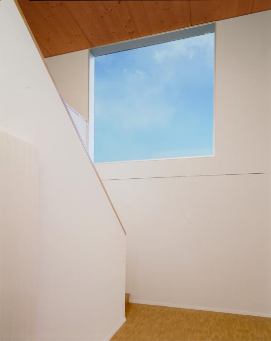 A staircase rising above the entryway to a building is painted with white and brown handrails and has square windows made of glass that lets in light from the skylight above.