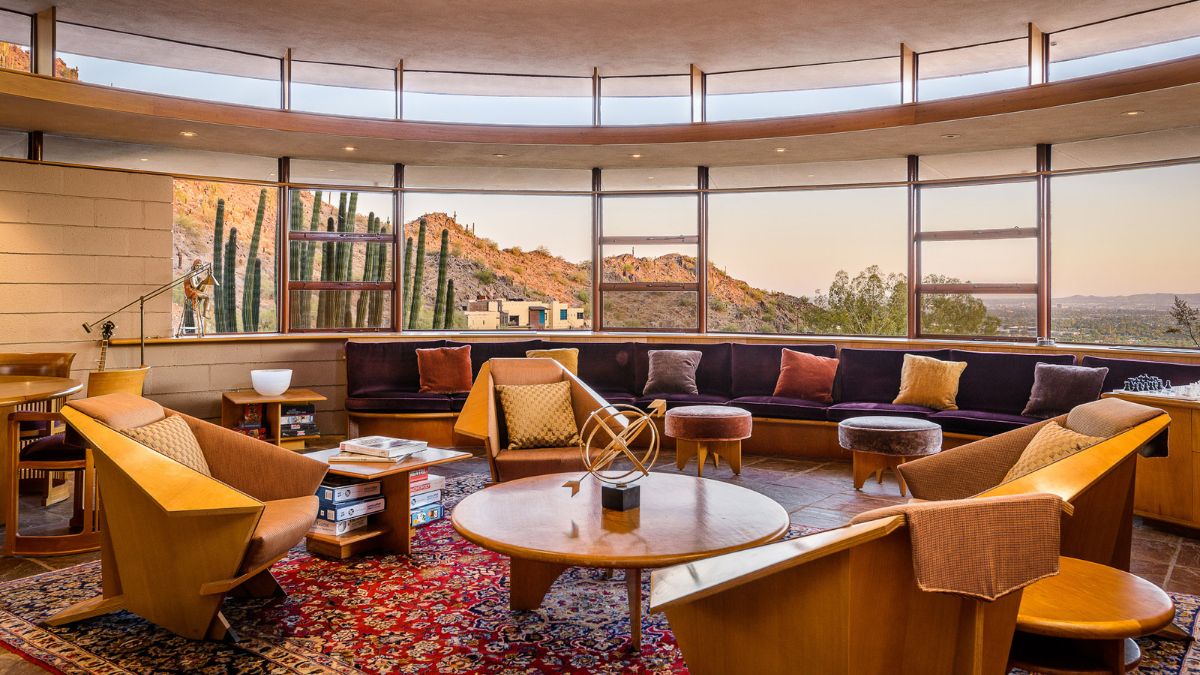 The living area is carpeted, has a high ceiling, and wooden furniture such as chairs and tables. It also includes large windows that overlook the mountain.