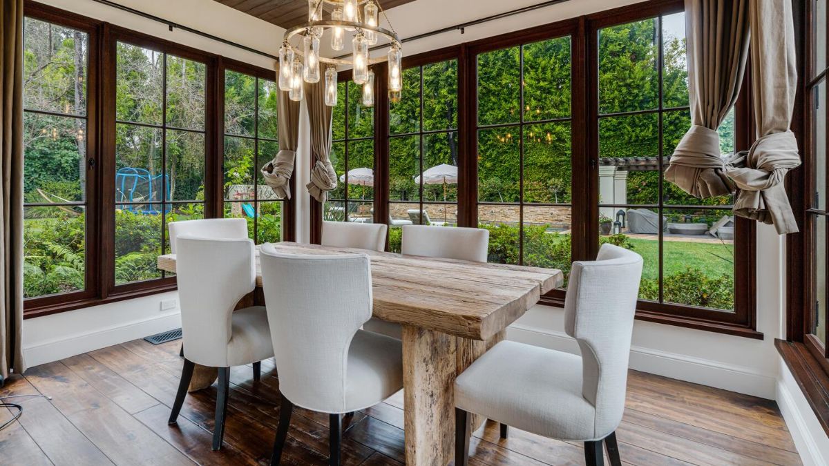 The dining area, which has hardwood floors, a wooden table with white chairs, and large windows that allow for a view of the outside, the chandelier further enhances the dining area's elegance.