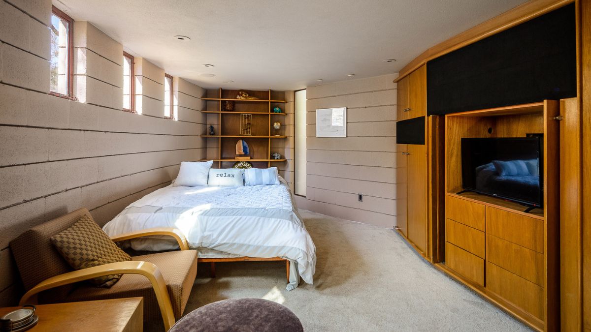 Carpeted bedroom with cemented white walls, wooden cupboards, wooden tiny table and chair with cushions, and square-shaped windows that provide light to the space.