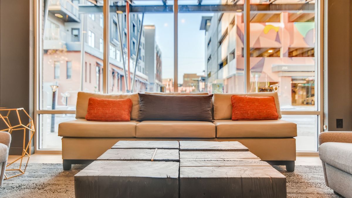 A large glass window behind an inviting orange and brown sofa in the hotel's living room area allows guests to see the nearby buildings.