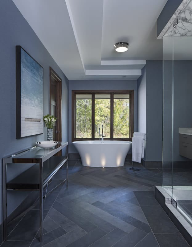 Grayish blue walls surround the primary bathroom. It has a freestanding tub brightened by wooden framed windows.