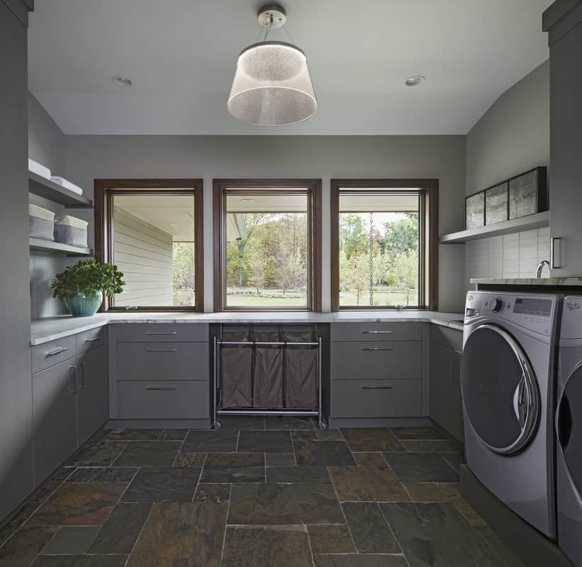 The laundry room has gray cabinets, floating shelves, and limestone flooring.