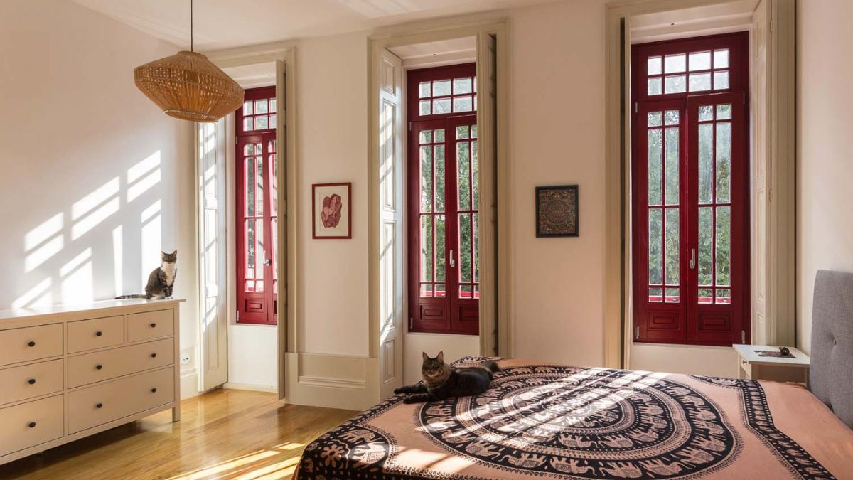 The bedroom has a beige color scheme, three wooden-glass windows, a local lamp, wood floors and a cabinet, as well as a bed with a distinctive design.