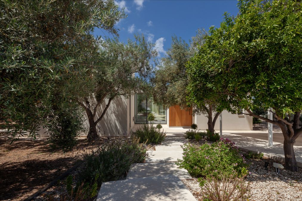 This is a photograph taken on a sunny day, depicting the dwelling encircled by trees and bushes, with a clear view of the pathway leading to the primary entrance of the residence.