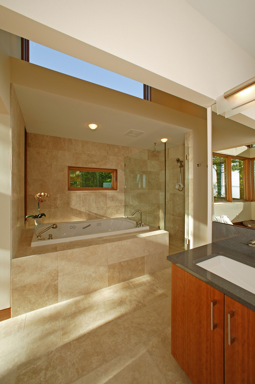 The bathroom features a deep soaking tub, a walk-in shower, travertine flooring, wood cabinetry, and an undermount sink with a full-width mirror.