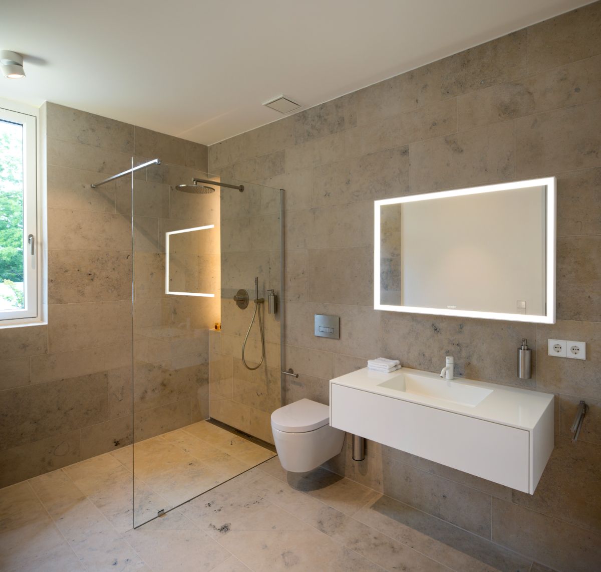 The bathroom also contains a walk-in shower with glass walls, a wall-mounted toilet, a floating sink, and an exposed concrete wall panel.