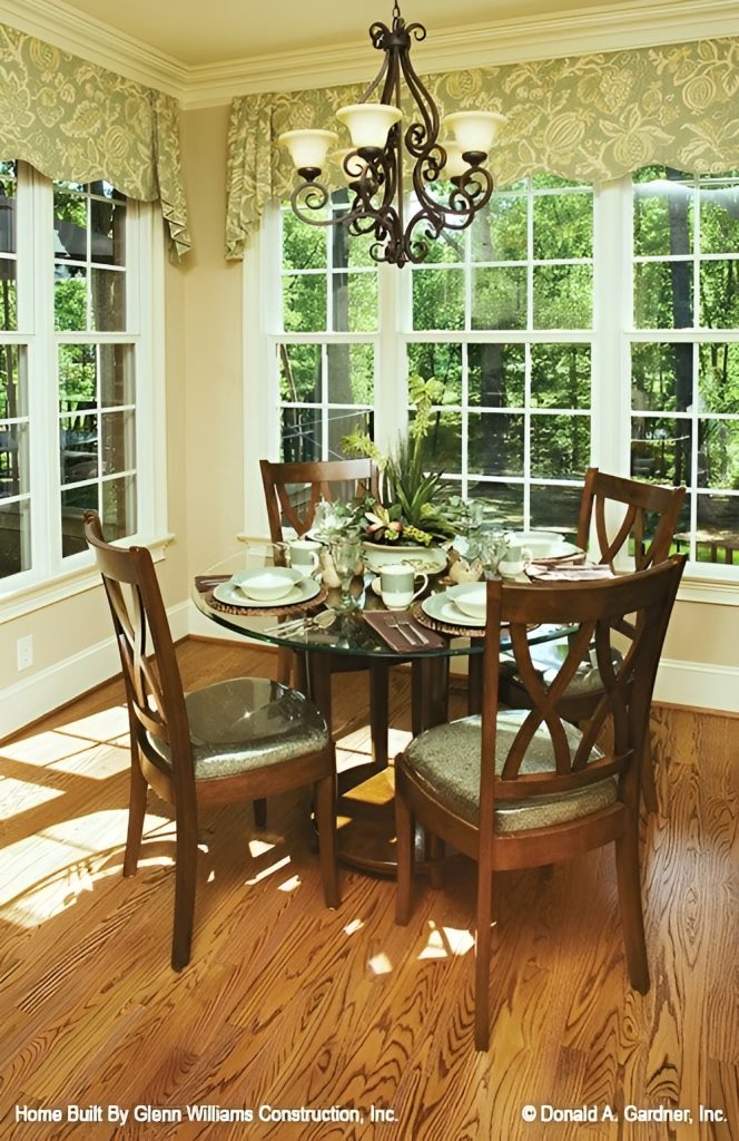 Breakfast nook with cushioned chairs and a round glass top table well-lit by an ornate chandelier.