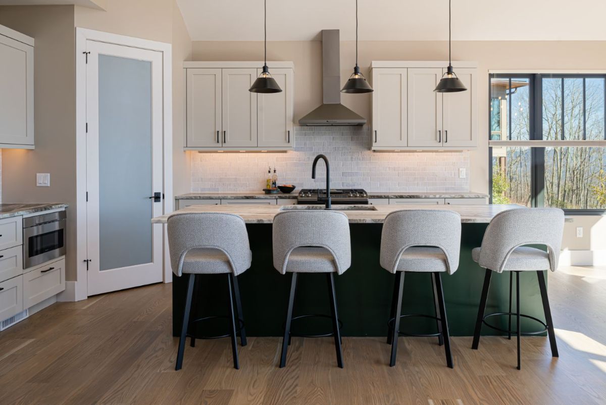 There is natural wood flooring in the kitchen, large windows, and built-in appliances. The dark green island stands out against the warm neutral color palette of the walls and ceiling.