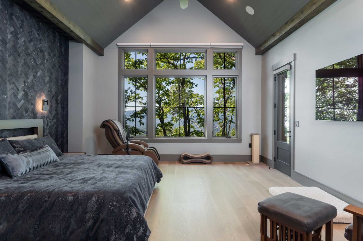 The vaulted ceiling, natural wood floors, and herringbone bed backdrop distinguish the main bedroom. The large windows let in plenty of natural light and provide a glimpse of the surrounding landscape.