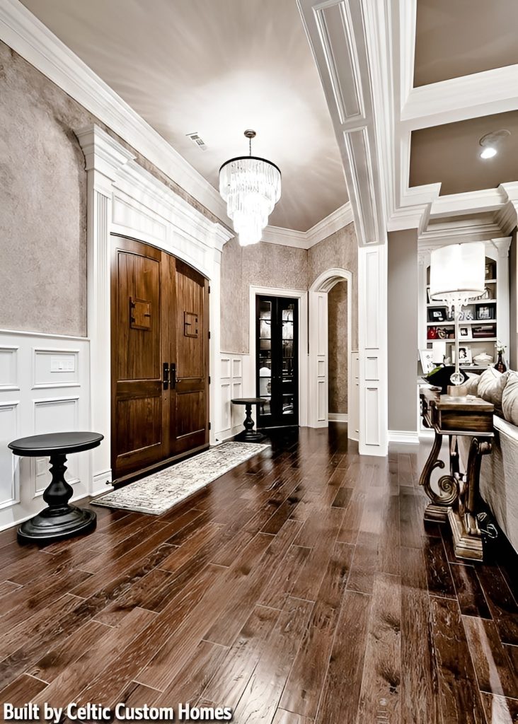 The home's foyer with a wooden front double door, a cascading chandelier, and a vintage runner flanked by black round tables.