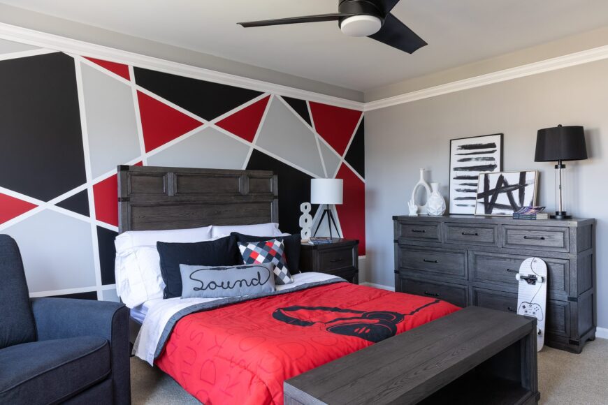 This wooden-framed bed takes center stage in this captivating bedroom, exuding warmth and elegance. The backdrop of the room features a striking geometric patterned wall, adorned in a tasteful combination of red, black, and gray hues. The wooden drawer cabinet with a harmonious display of ceramic pots, art pieces, and a lampshade adds a touch of practicality and natural charm to the overall aesthetic.