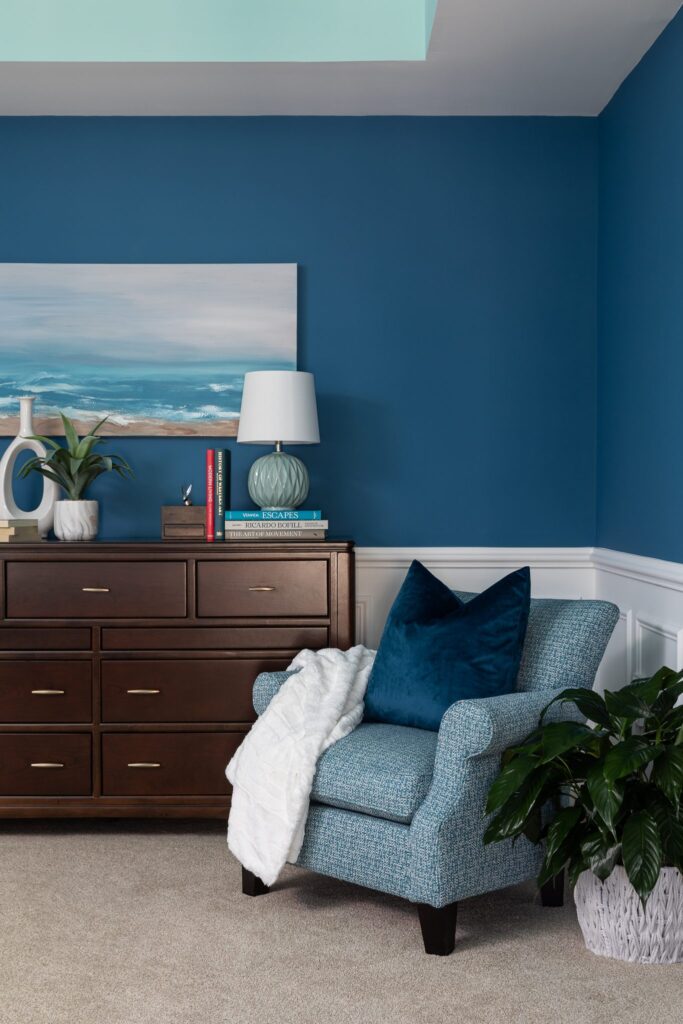 The midnight blue colored wall captures attention with its serene charm. It is adorned with an artful depiction of sea waves, bringing a touch of the ocean indoors. The wooden drawer cabinet and the cozy midnight blue sofa chair nestled in the corner complement the wall, adding a rustic touch to the scene.