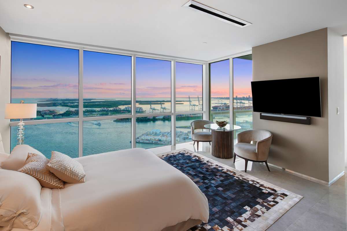 A bedroom with a TV, chairs, and a coffee table, surrounded by expansive glass windows