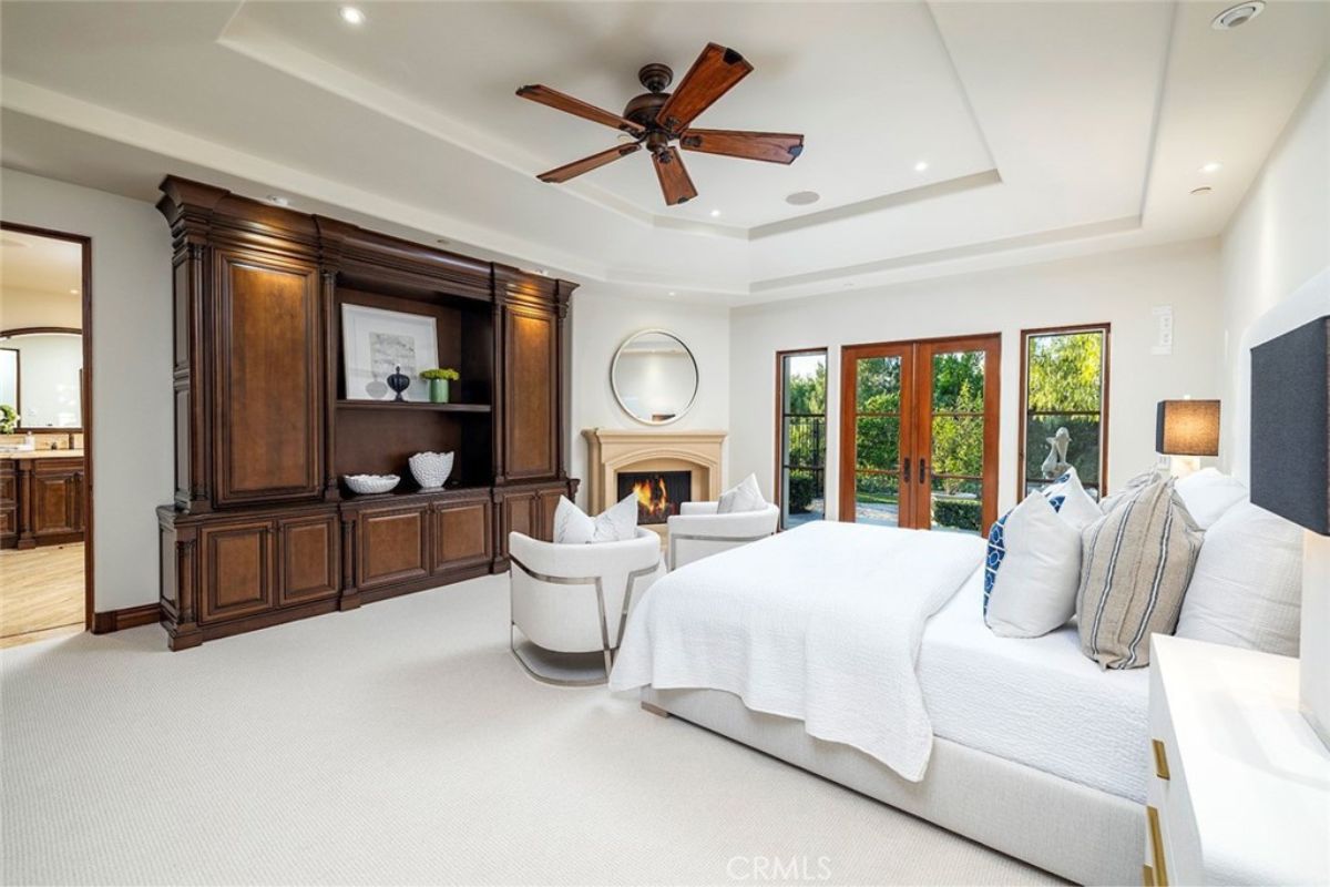 A bedroom with a white bed, walls, ceiling, fireplace, and ceiling fan.