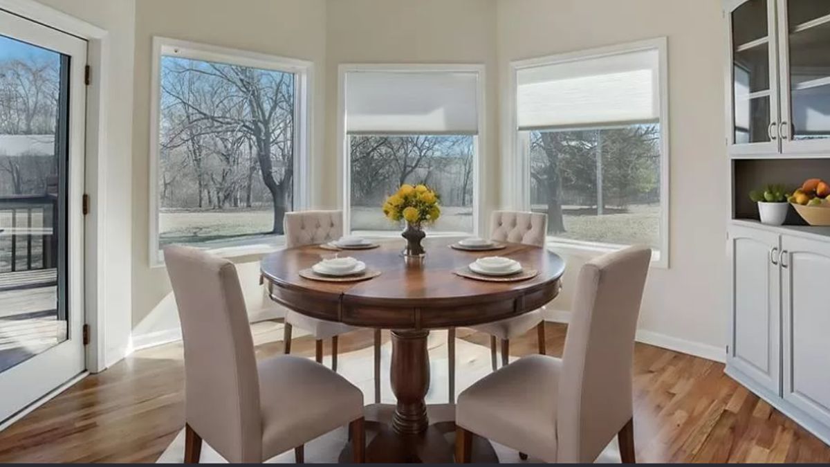 A breakfast nook with windows and a round table surrounded by chairs.