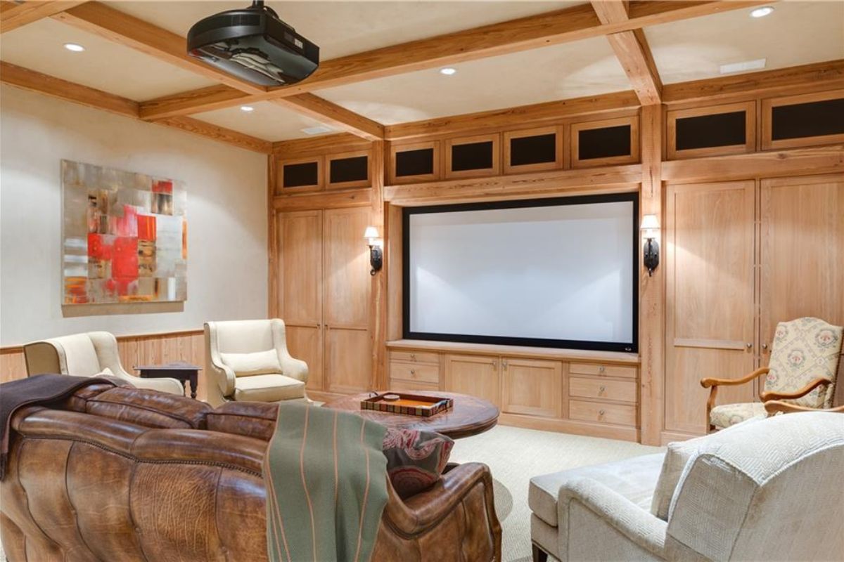 A home theater with an exposed beam ceiling, paintings, a large screen, drawers, couches, chairs, and a center table.