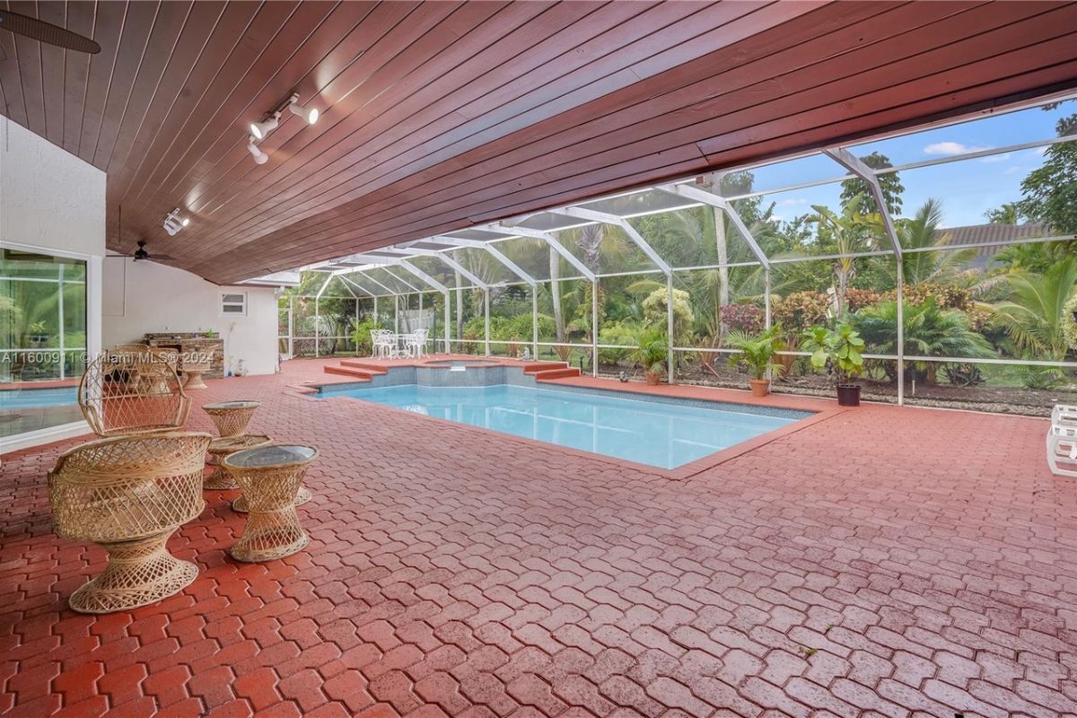 A covered swimming pool with chairs, a coffee table, plants, glass walls, and a glass ceiling.