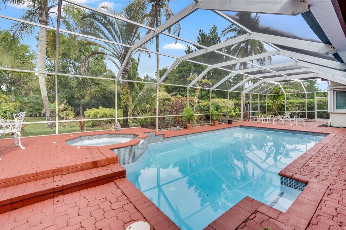 A covered swimming pool with chairs, a coffee table, plants, glass walls, and a glass ceiling.