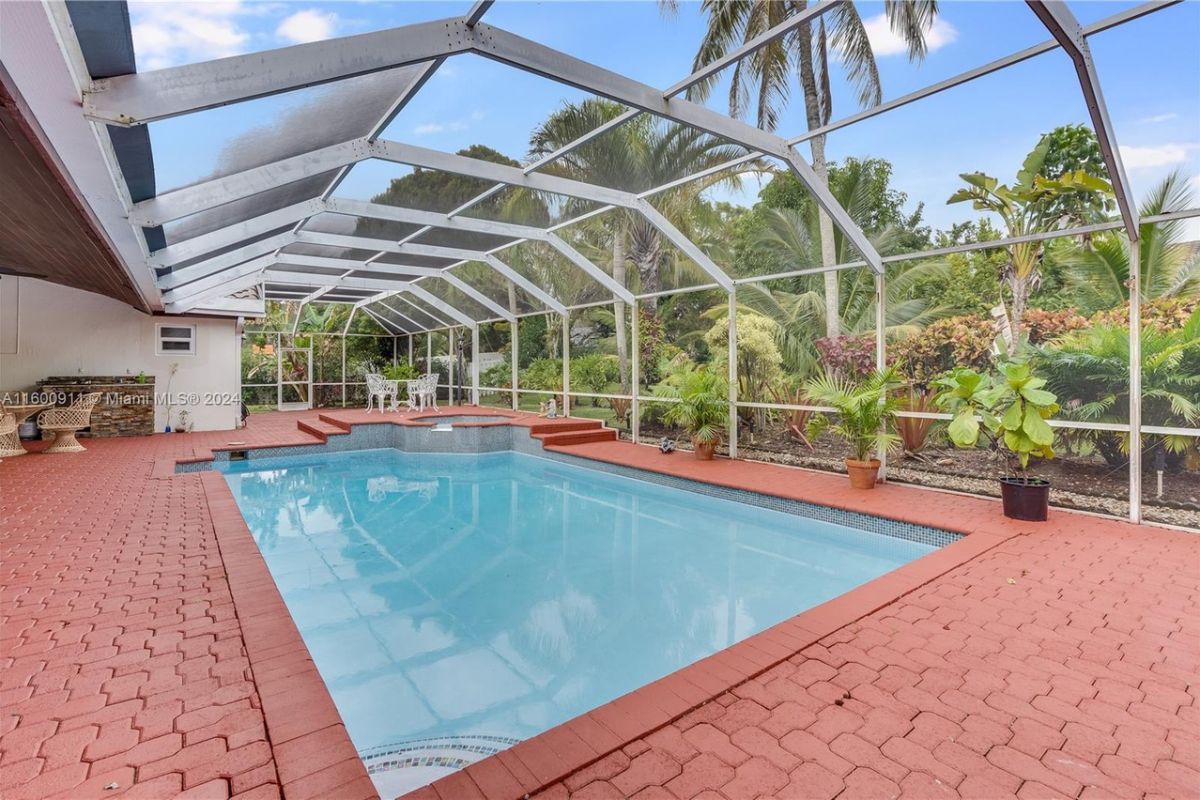 A covered swimming pool with chairs, a coffee table, plants, glass walls, and a glass ceiling.