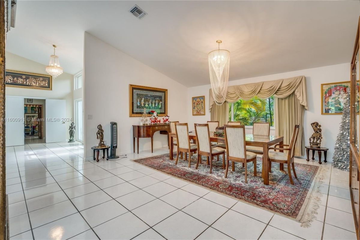 A dining room with a tiled floor, paintings, windows, a rug, and a dining table surrounded by chairs.