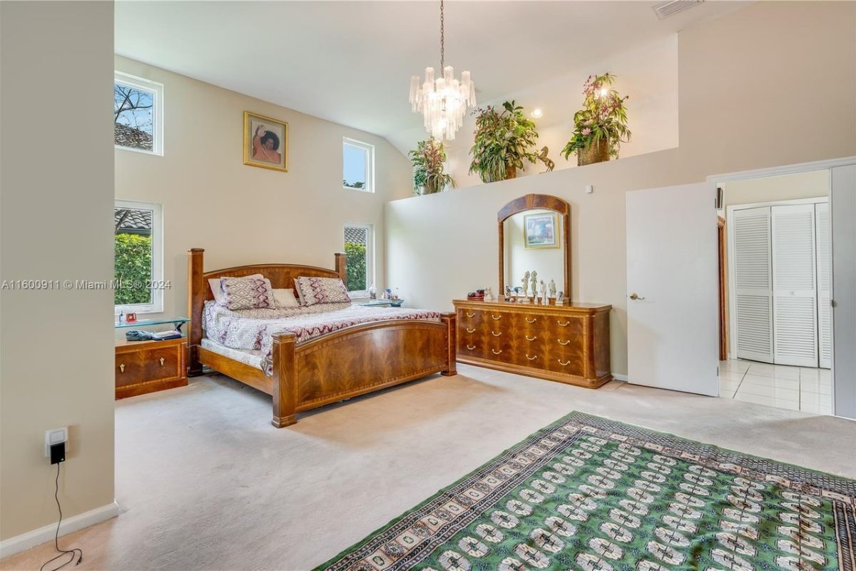 A bedroom with a bed, high ceiling, chandelier, windows, mirror, and cabinets.