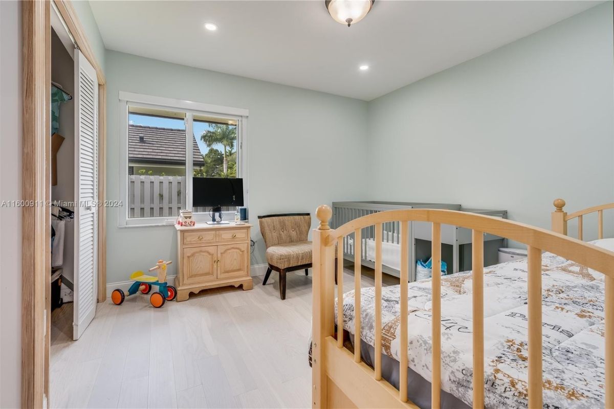 A bedroom with a bed, window, chair, crib, and cabinet.