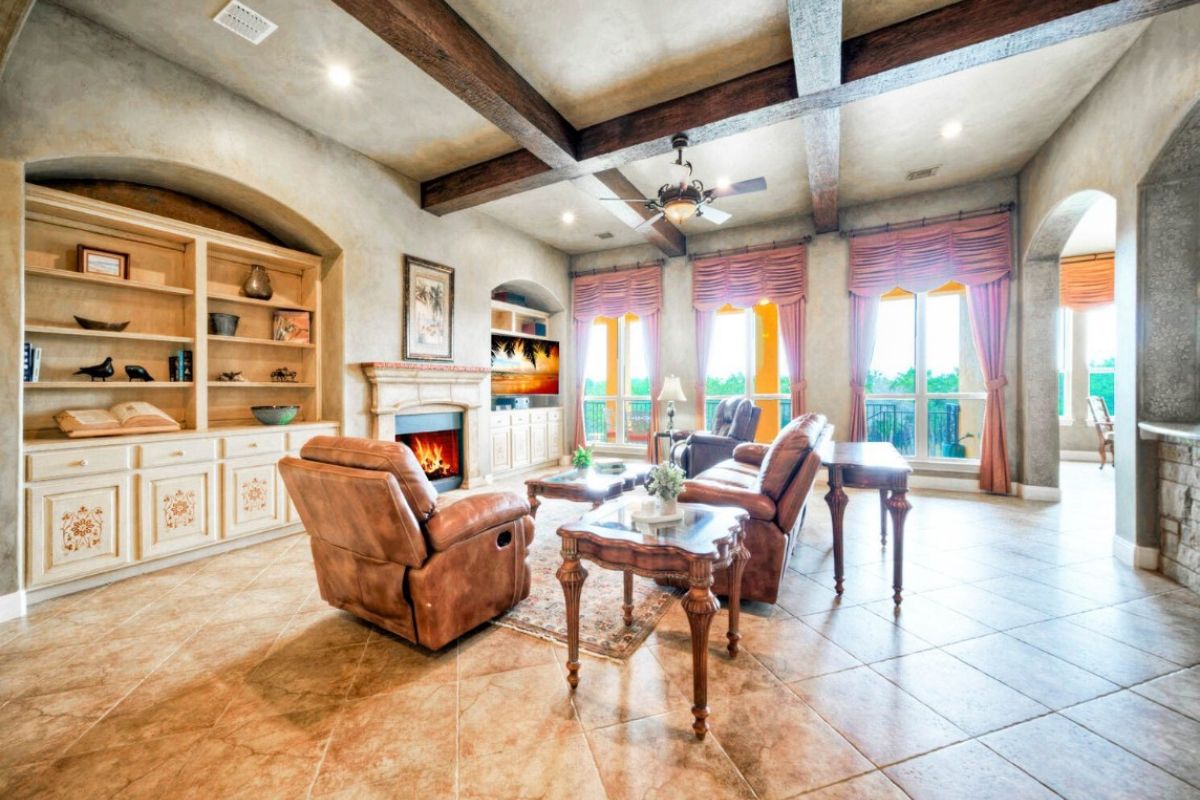 A living room with an exposed beam ceiling, tiled floor, shelf, cabinet, windows, couch, chairs, and a center table.