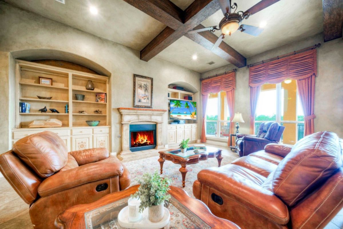 A living room with an exposed beam ceiling, fireplce, shelf, cabinet, windows, couch, chairs, and a center table.