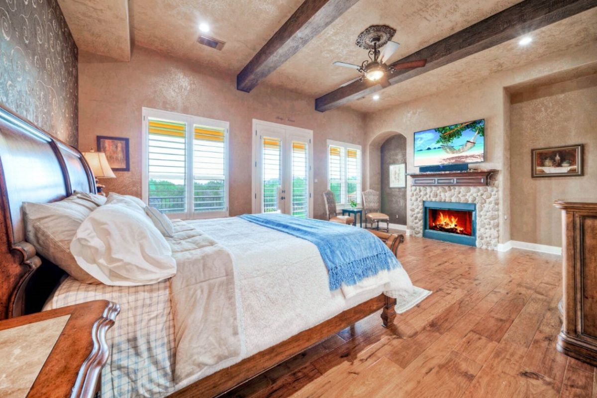 A bedroom with a bed, exposed beam ceiling, fireplace, TV, and window.